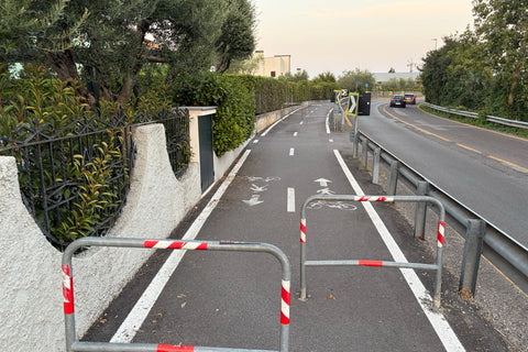 A shared cycle and pedestrian path beside a road near Lake Garda, blocked by pairs of red and white metal barriers